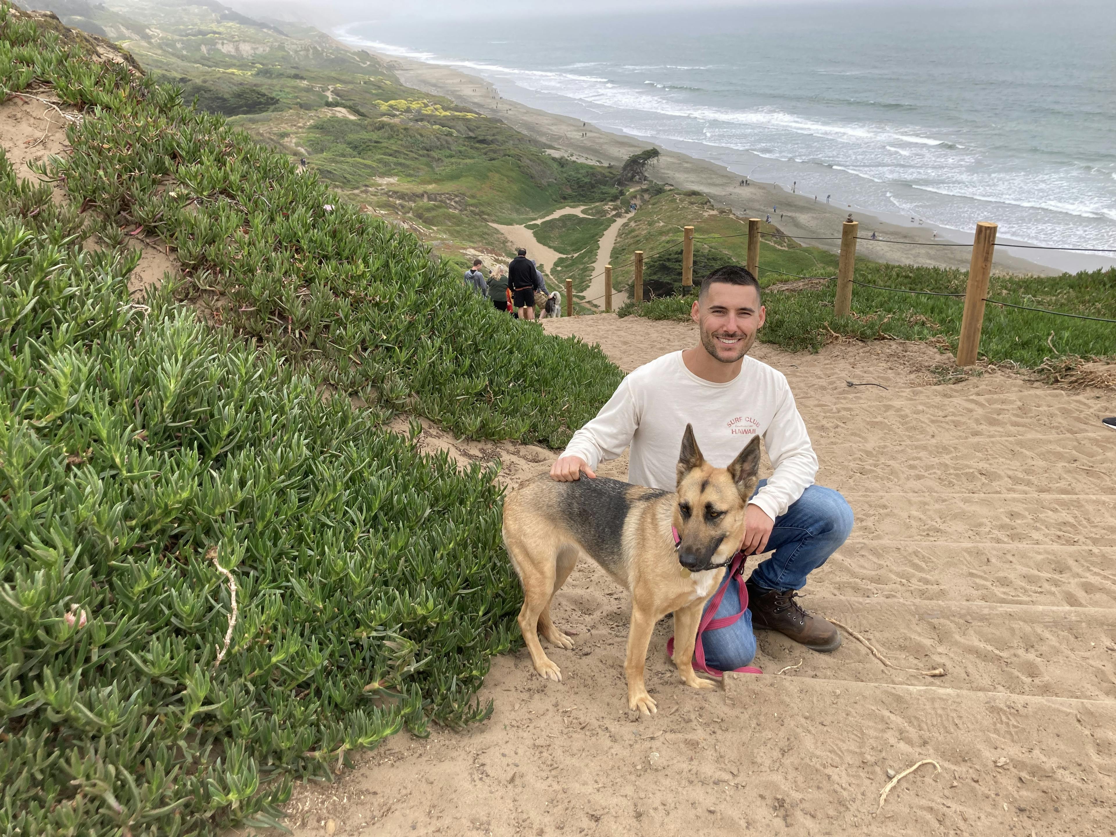 A photo of Nico petting his German Shepherd Sophie on a sandy hill overlooking the ocean at Fort Funston in San Francisco.