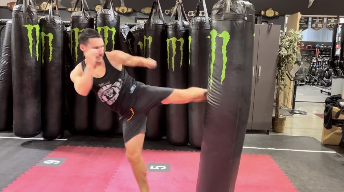 A photo of Nico kicking a heavy bag with a Muay Thai round kick.