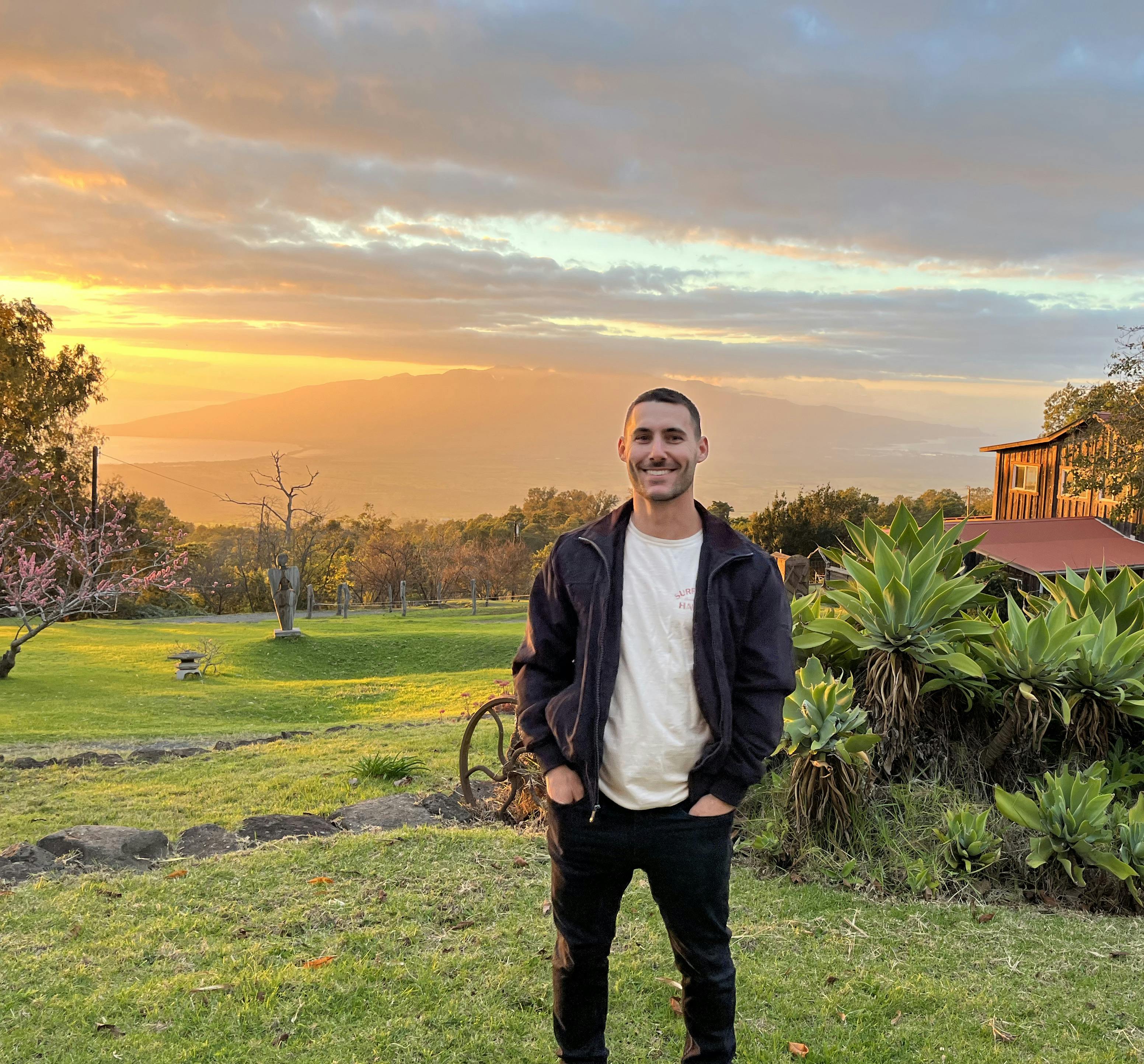 A photo of Nico standing in a lush meadow overlooking the West Maui Mountains as the sun sets.