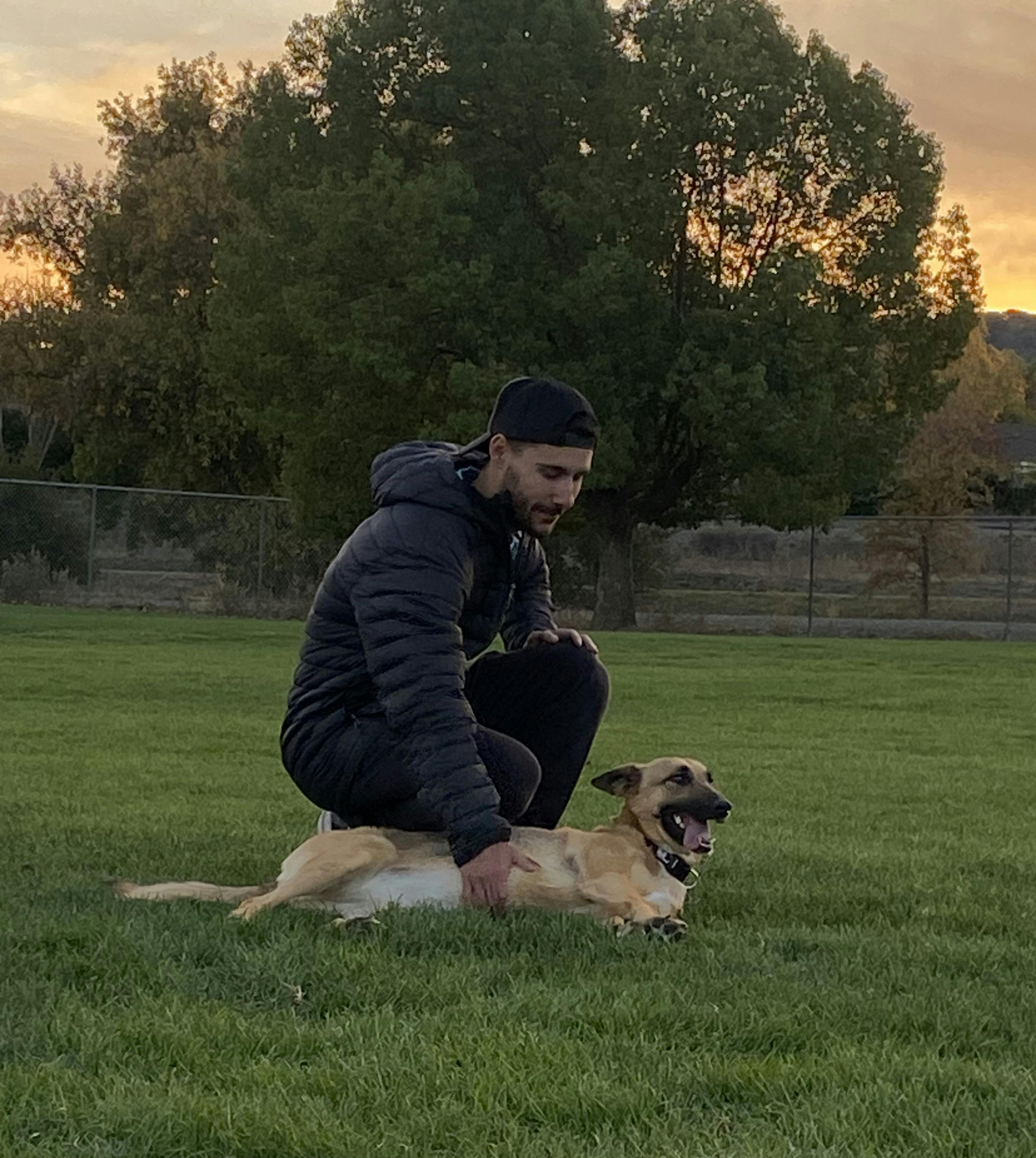 A photo of Nico petting his German Shepherd Sophie's belly while she lays on the grass at the park as the sun sets.